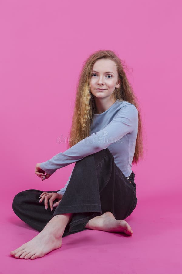 Teenage Girl In Casual Street Clothing Sitting With Legs Folded Against Pink Background