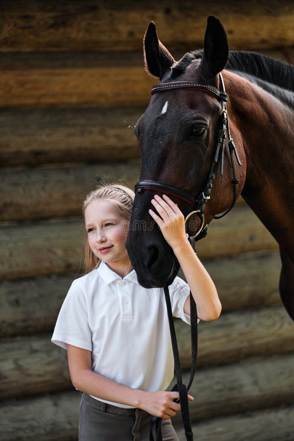 A Teenage Girl Jockey Stands Next To A Brown Horse And Hugs Her. Against The Background Of A Wooden Wall Stables.