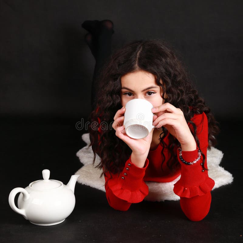 Teenage girl in red blouse and black pantyhose drinking tea from white cup while lying on white sheep fur. Teapot aside of her. Black background. Tea party concept. Teenage girl in red blouse and black pantyhose drinking tea from white cup while lying on white sheep fur. Teapot aside of her. Black background. Tea party concept.