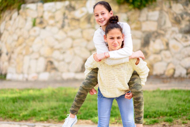 Teenage Sisters On A City Park In The Summertime. Two Young Girls Playing A Game Together At Sunny Day. Best Friends Having Fun In The Summer Riding each other`s back. Teenage Sisters On A City Park In The Summertime. Two Young Girls Playing A Game Together At Sunny Day. Best Friends Having Fun In The Summer Riding each other`s back.