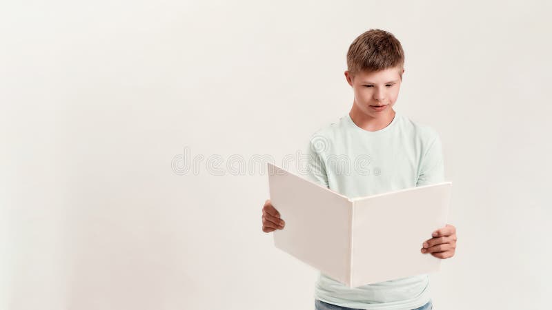 Teenaged disabled boy with Down syndrome looking focused while reading a book, standing isolated over white background