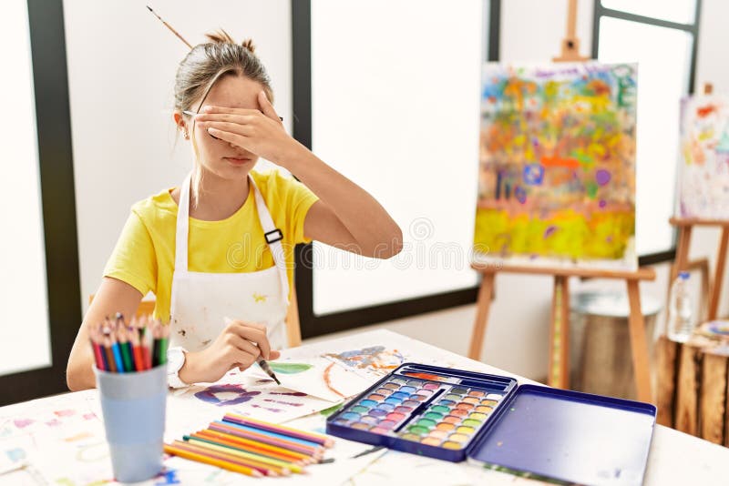 Young brunette teenager at art studio covering eyes with hand, looking serious and sad. sightless, hiding and rejection concept. Young brunette teenager at art studio covering eyes with hand, looking serious and sad. sightless, hiding and rejection concept