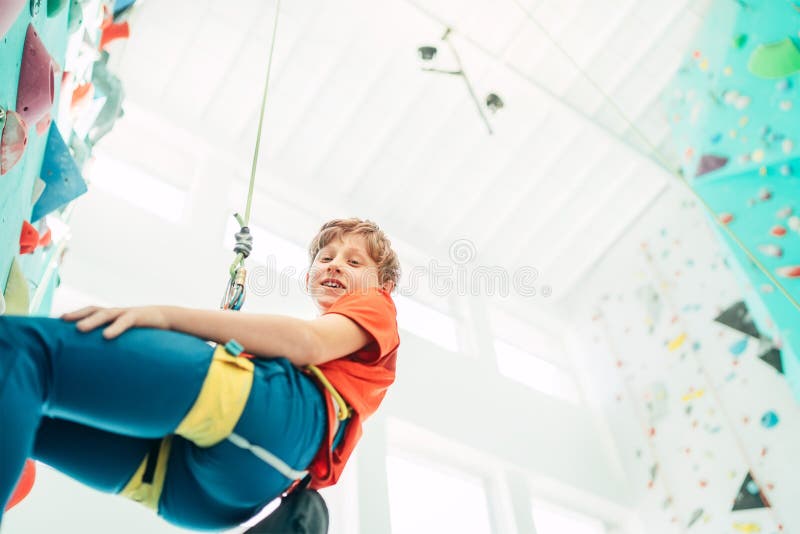 Teenager boy at indoor climbing wall hall. Boy is climbing using a top rope and climbing harness and somebody belaying him from fl