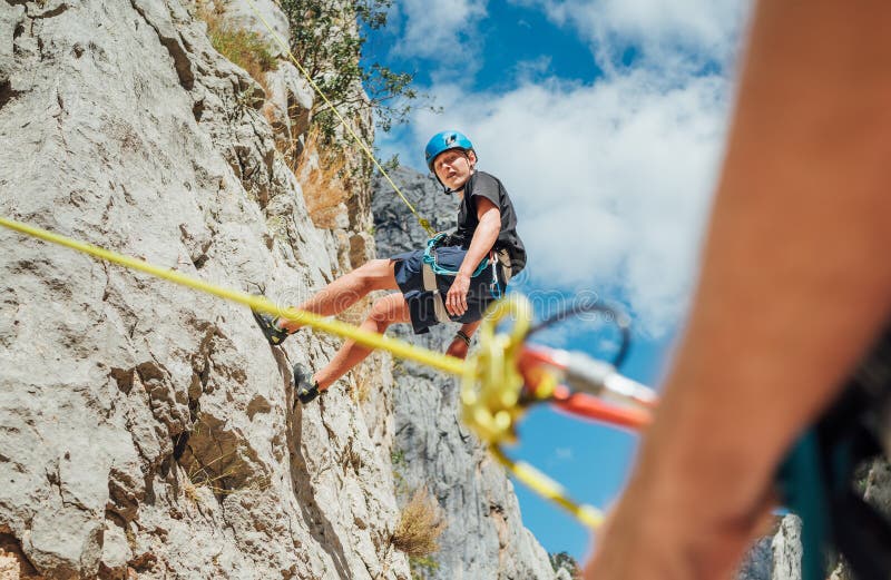 Teenager boy on the natural cliff climbing wall. Boy hanging on a rope using a climbing harness and the father belaying him on the