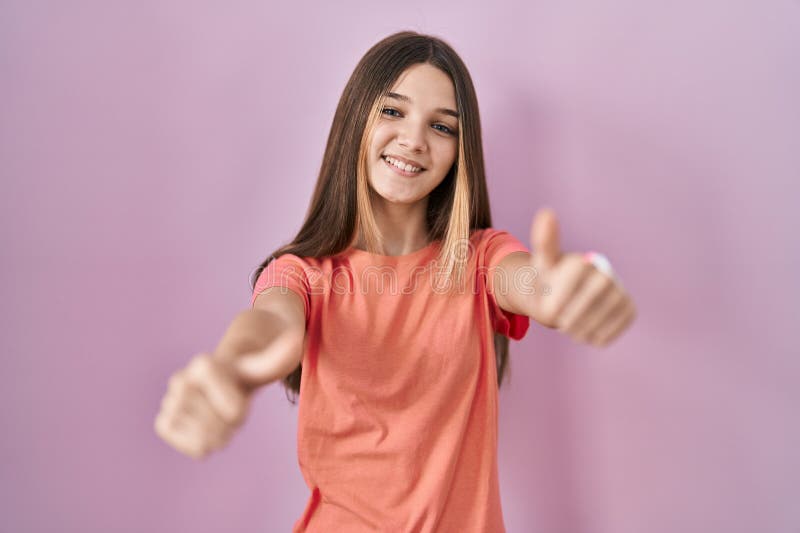 Teenager girl standing over pink background approving doing positive gesture with hand, thumbs up smiling and happy for success