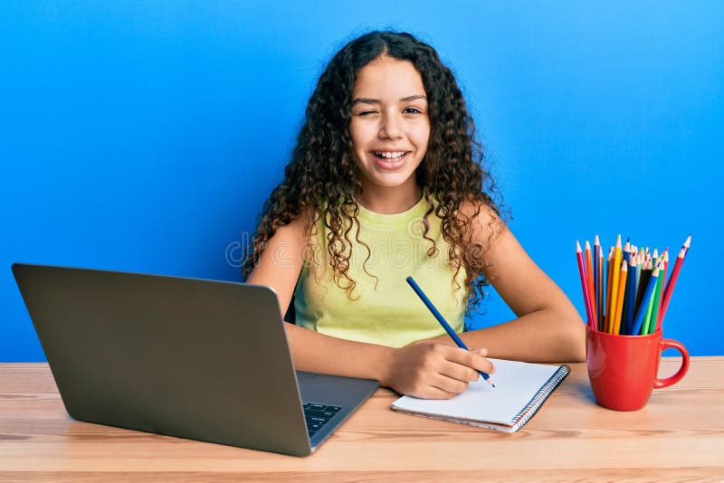 Teenager hispanic girl sitting on the table studying for school winking looking at the camera with sexy expression, cheerful and happy face