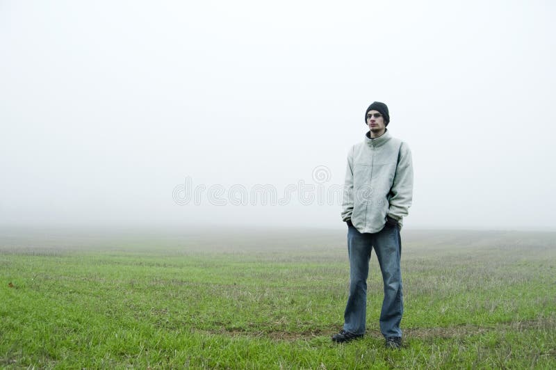 Biela kaukazský teenager stojí v zelenej tráve oblasti čiernu čiapočku a biela bunda s jeho rúk v jeho modré džínsy.