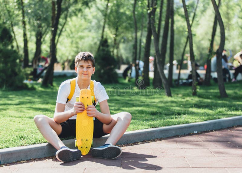 Teenager in white t shirt spends free time training skating in the city park. Boy is looking at camera.