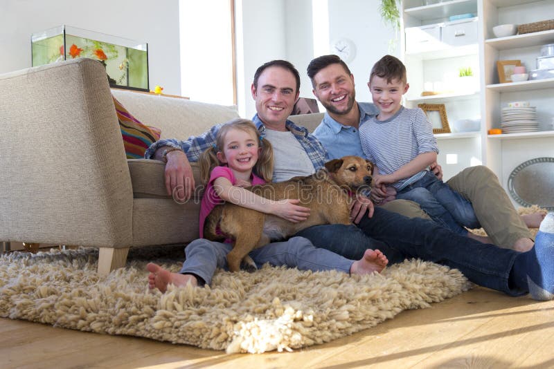 Same sex male couple sitting on the floor in their living room with their son and daughter. Their pet dog is lying across them. Same sex male couple sitting on the floor in their living room with their son and daughter. Their pet dog is lying across them.