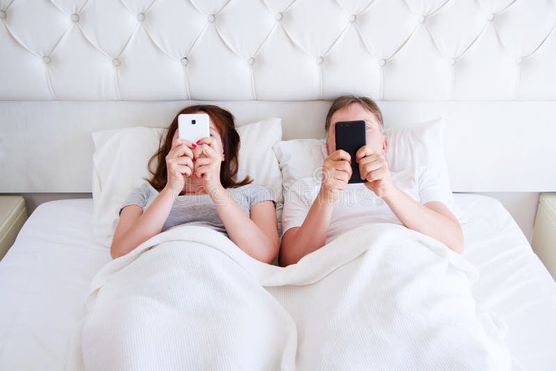 Telefoni cellulari invecchiati medi in camera da letto, concetto moderno della tenuta delle coppie della gente immagini stock Telefoni cellulari invecchiati medi in camera da letto, concetto moderno della tenuta delle coppie della gente immagini stock