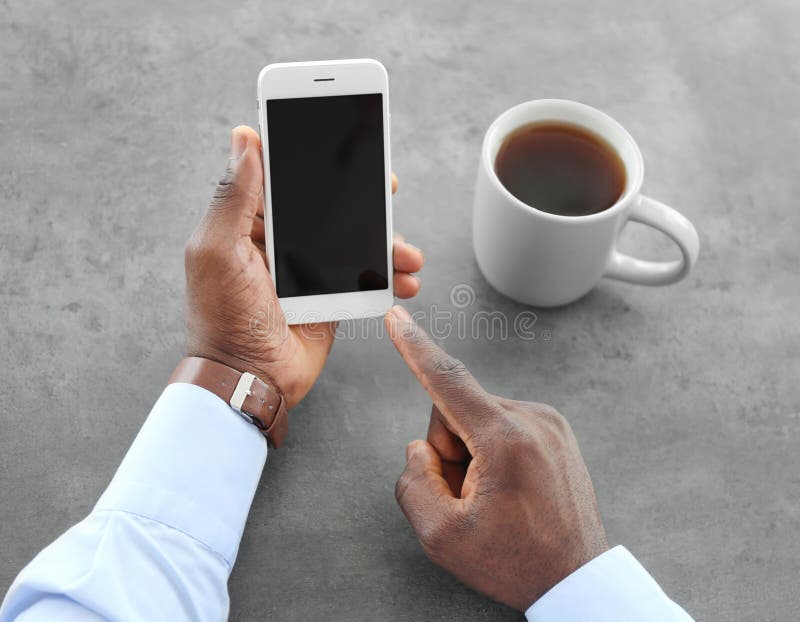 Telefono afroamericano della tenuta dell'uomo con lo schermo in bianco a disposizione alla tavola fotografie stock Telefono afroamericano della tenuta dell'uomo con lo schermo in bianco a disposizione alla tavola fotografie stock