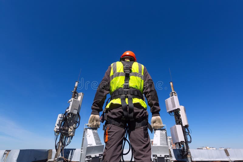 Telecommunication engineer in helmet and uniform holds telecomunication equipment in his hand and antennas of GSM DCS UMTS LTE bands, outdoor radio units, optic fibers, power cables are installed on the roof. Working process of upgrading telecommunication equipment. Telecommunication engineer in helmet and uniform holds telecomunication equipment in his hand and antennas of GSM DCS UMTS LTE bands, outdoor radio units, optic fibers, power cables are installed on the roof. Working process of upgrading telecommunication equipment.