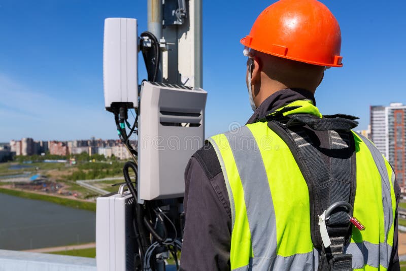 Telecommunication engineer in helmet and uniform holds telecomunication equipment in his hand and antennas of GSM DCS UMTS LTE bands, outdoor radio units, optic fibers, power cables are installed on the roof. Working process of upgrading telecommunication equipment. Telecommunication engineer in helmet and uniform holds telecomunication equipment in his hand and antennas of GSM DCS UMTS LTE bands, outdoor radio units, optic fibers, power cables are installed on the roof. Working process of upgrading telecommunication equipment.