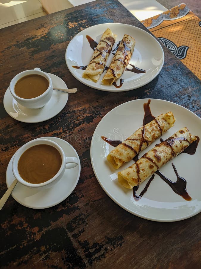 Zanzibar, Tanzania - December 10, 2019: 2 plates of pancakes and 2 cups of coffee. Pancakes with chocolate on the plate. Coffee with milk is poured in cups. Vertical. Zanzibar, Tanzania - December 10, 2019: 2 plates of pancakes and 2 cups of coffee. Pancakes with chocolate on the plate. Coffee with milk is poured in cups. Vertical