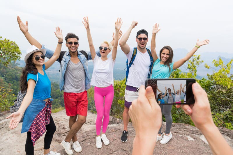 Teléfono elegante de la célula que toma la foto del grupo turístico alegre con la mochila sobre paisaje del top de la montaña, pr foto de archivo Teléfono elegante de la célula que toma la foto del grupo turístico alegre con la mochila sobre paisaje del top de la montaña, pr foto de archivo