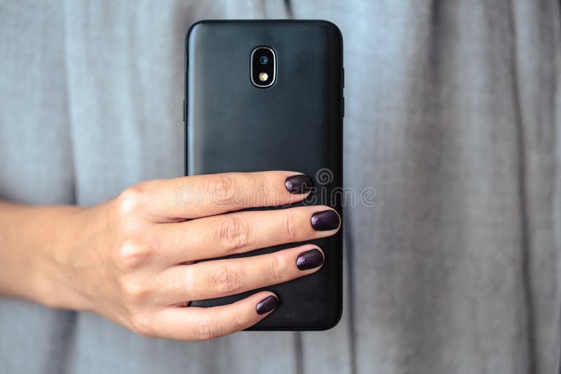 Black mobie smart phone in woman/s hands with perfect dark violet manicure over simple background. Black mobie smart phone in woman/s hands with perfect dark violet manicure over simple background