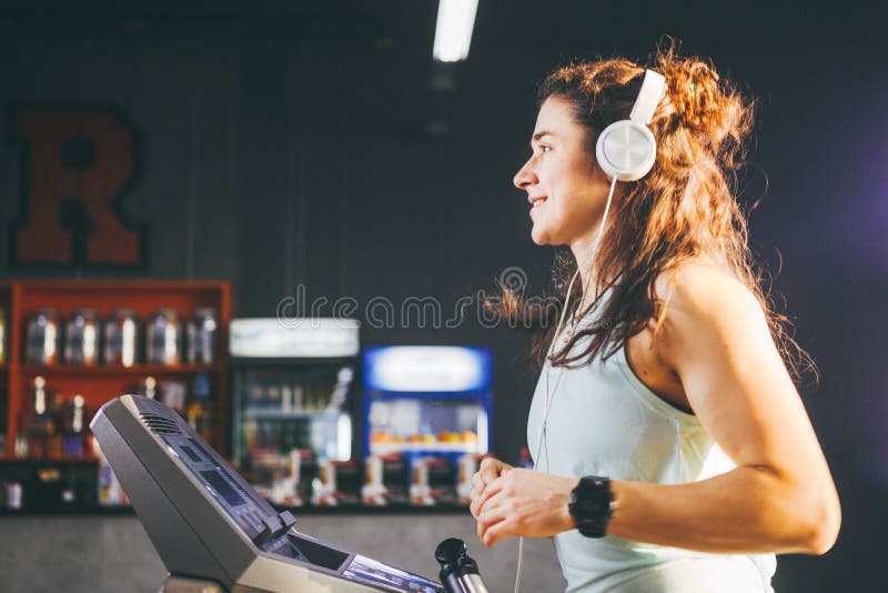 Temat är sporten och musik En härlig uppblåst kvinna kör i idrottshallen på en trampkvarn På hennes huvud är den stora vita hörlu arkivbild Temat är sporten och musik En härlig uppblåst kvinna kör i idrottshallen på en trampkvarn På hennes huvud är den stora vita hörlu arkivbild