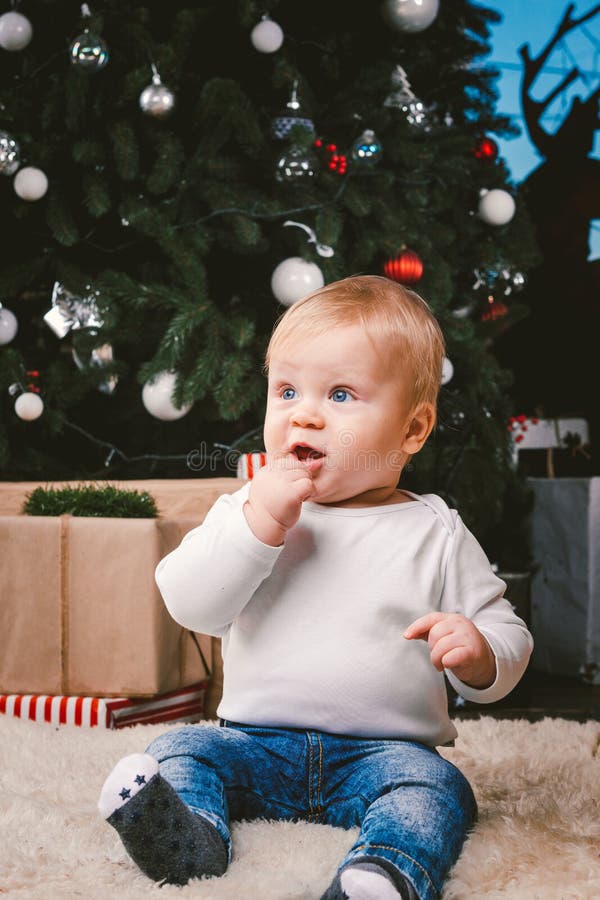 Theme winter and Christmas holidays. Child boy Caucasian blond 1 year old sitting home floor near Christmas tree with New Year decor on shaggy carpet skin receives gifts, opens gift boxes in evening. Theme winter and Christmas holidays. Child boy Caucasian blond 1 year old sitting home floor near Christmas tree with New Year decor on shaggy carpet skin receives gifts, opens gift boxes in evening.