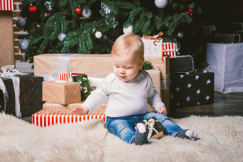 Theme winter and Christmas holidays. Child boy Caucasian blond 1 year old sitting home floor near Christmas tree with New Year decor on shaggy carpet skin receives gifts, opens gift boxes in evening. Theme winter and Christmas holidays. Child boy Caucasian blond 1 year old sitting home floor near Christmas tree with New Year decor on shaggy carpet skin receives gifts, opens gift boxes in evening.