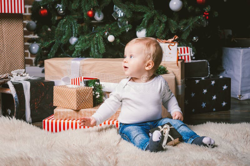 Theme winter and Christmas holidays. Child boy Caucasian blond 1 year old sitting home floor near Christmas tree with New Year decor on shaggy carpet skin receives gifts, opens gift boxes in evening. Theme winter and Christmas holidays. Child boy Caucasian blond 1 year old sitting home floor near Christmas tree with New Year decor on shaggy carpet skin receives gifts, opens gift boxes in evening.