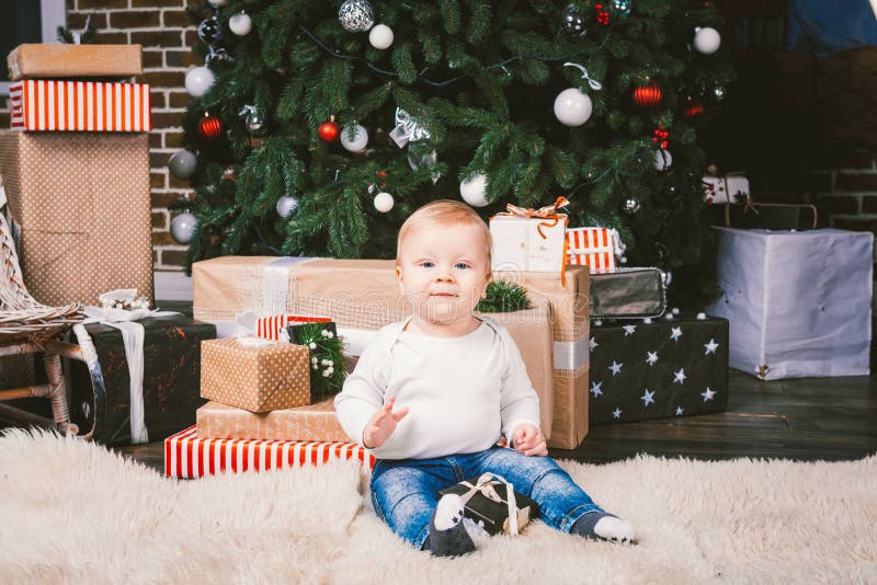 Theme winter and Christmas holidays. Child boy Caucasian blond 1 year old sitting home floor near Christmas tree with New Year decor on shaggy carpet skin receives gifts, opens gift boxes in evening. Theme winter and Christmas holidays. Child boy Caucasian blond 1 year old sitting home floor near Christmas tree with New Year decor on shaggy carpet skin receives gifts, opens gift boxes in evening.