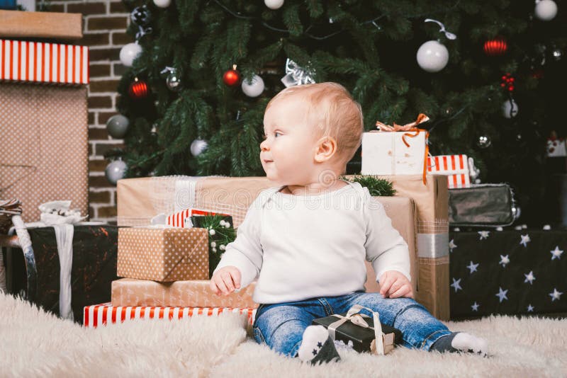 Theme winter and Christmas holidays. Child boy Caucasian blond 1 year old sitting home floor near Christmas tree with New Year decor on shaggy carpet skin receives gifts, opens gift boxes in evening. Theme winter and Christmas holidays. Child boy Caucasian blond 1 year old sitting home floor near Christmas tree with New Year decor on shaggy carpet skin receives gifts, opens gift boxes in evening.