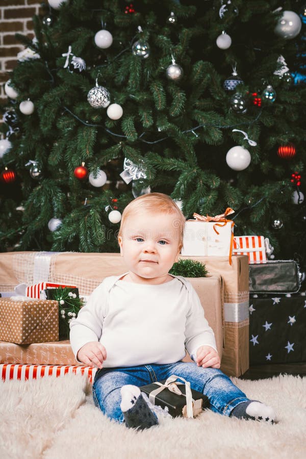 Theme winter and Christmas holidays. Child boy Caucasian blond 1 year old sitting home floor near Christmas tree with New Year decor on shaggy carpet skin receives gifts, opens gift boxes in evening. Theme winter and Christmas holidays. Child boy Caucasian blond 1 year old sitting home floor near Christmas tree with New Year decor on shaggy carpet skin receives gifts, opens gift boxes in evening.