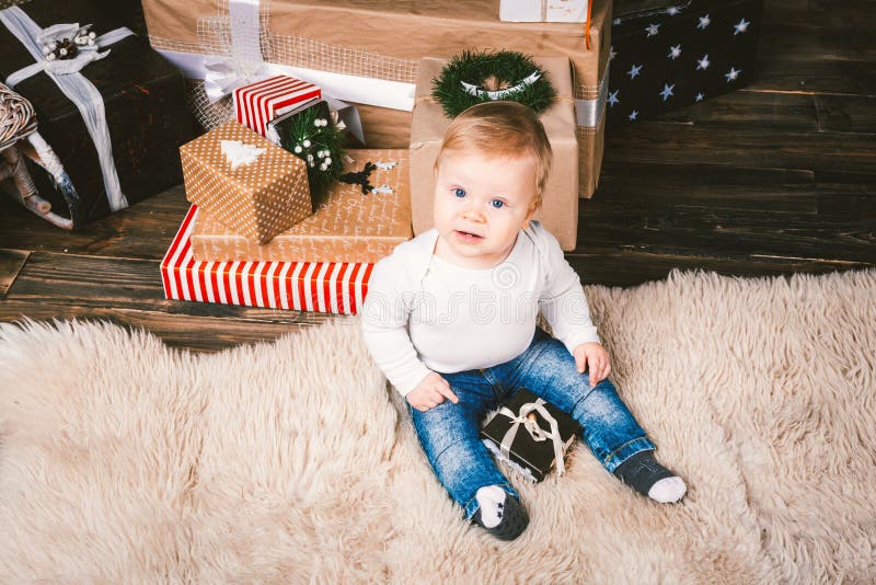 Theme winter and Christmas holidays. Child boy Caucasian blond 1 year old sitting home floor near Christmas tree with New Year decor on shaggy carpet skin receives gifts, opens gift boxes in evening. Theme winter and Christmas holidays. Child boy Caucasian blond 1 year old sitting home floor near Christmas tree with New Year decor on shaggy carpet skin receives gifts, opens gift boxes in evening.