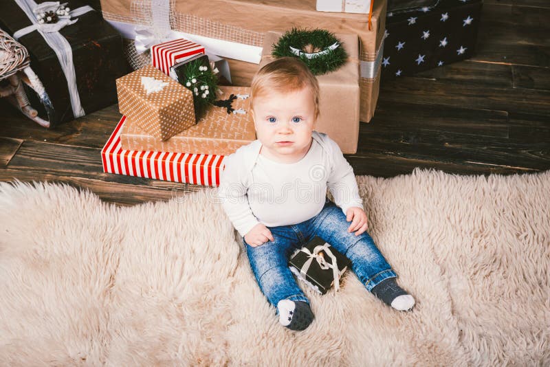 Theme winter and Christmas holidays. Child boy Caucasian blond 1 year old sitting home floor near Christmas tree with New Year decor on shaggy carpet skin receives gifts, opens gift boxes in evening. Theme winter and Christmas holidays. Child boy Caucasian blond 1 year old sitting home floor near Christmas tree with New Year decor on shaggy carpet skin receives gifts, opens gift boxes in evening.