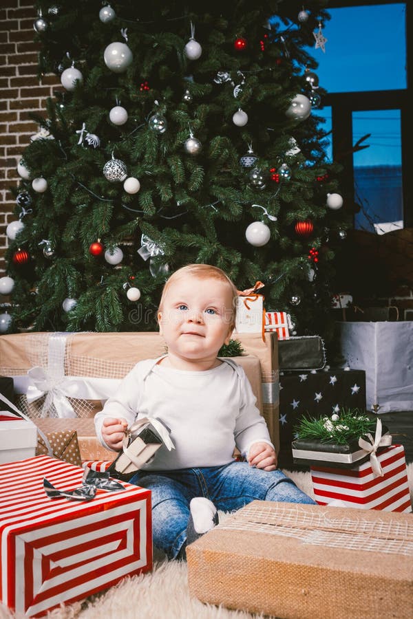 Theme winter and Christmas holidays. Child boy Caucasian blond 1 year old sitting home floor near Christmas tree with New Year decor on shaggy carpet skin receives gifts, opens gift boxes in evening. Theme winter and Christmas holidays. Child boy Caucasian blond 1 year old sitting home floor near Christmas tree with New Year decor on shaggy carpet skin receives gifts, opens gift boxes in evening.