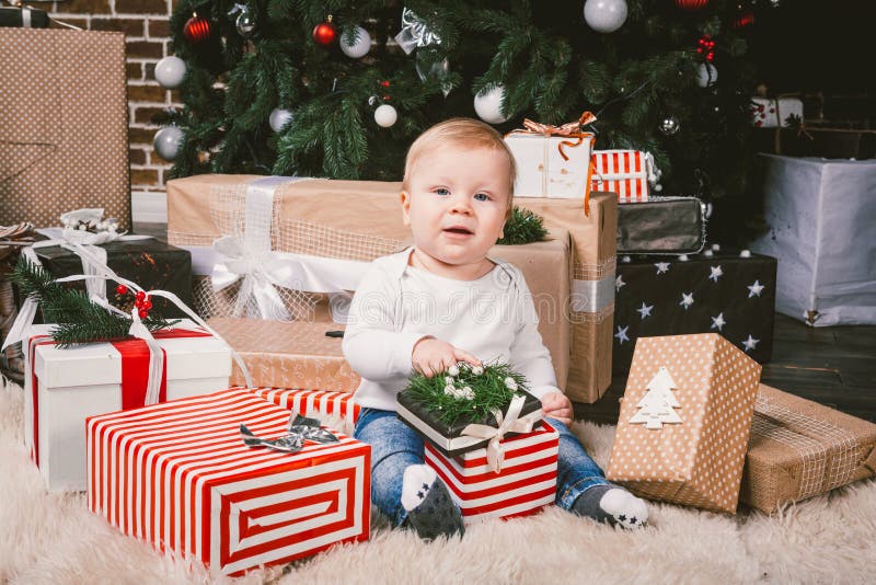 Theme winter and Christmas holidays. Child boy Caucasian blond 1 year old sitting home floor near Christmas tree with New Year decor on shaggy carpet skin receives gifts, opens gift boxes in evening. Theme winter and Christmas holidays. Child boy Caucasian blond 1 year old sitting home floor near Christmas tree with New Year decor on shaggy carpet skin receives gifts, opens gift boxes in evening.