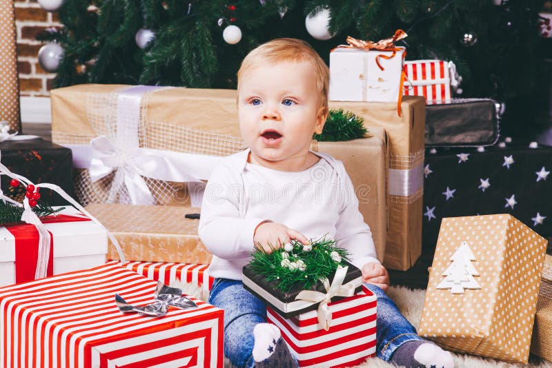 Theme winter and Christmas holidays. Child boy Caucasian blond 1 year old sitting home floor near Christmas tree with New Year decor on shaggy carpet skin receives gifts, opens gift boxes in evening. Theme winter and Christmas holidays. Child boy Caucasian blond 1 year old sitting home floor near Christmas tree with New Year decor on shaggy carpet skin receives gifts, opens gift boxes in evening.