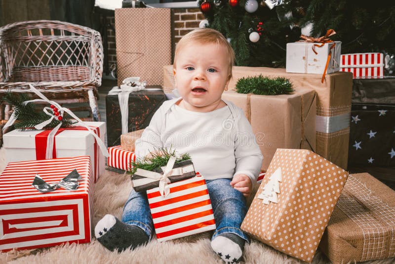 Theme winter and Christmas holidays. Child boy Caucasian blond 1 year old sitting home floor near Christmas tree with New Year decor on shaggy carpet skin receives gifts, opens gift boxes in evening. Theme winter and Christmas holidays. Child boy Caucasian blond 1 year old sitting home floor near Christmas tree with New Year decor on shaggy carpet skin receives gifts, opens gift boxes in evening.