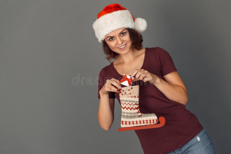 Tempo di natale Giovane donna in cappello di Santa sul regalo mettente grigio nel sorridere dello stivale del pattino felice immagine stock libera da diritti Tempo di natale Giovane donna in cappello di Santa sul regalo mettente grigio nel sorridere dello stivale del pattino felice immagine stock libera da diritti