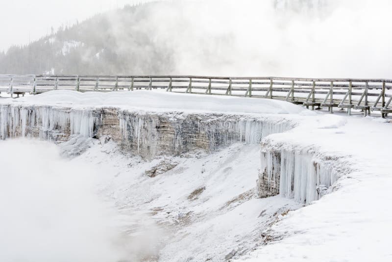 Curtain of icicles covering a layered wall of stone in front of a raised boardwalk and hilly, tree-covered terrain. Curtain of icicles covering a layered wall of stone in front of a raised boardwalk and hilly, tree-covered terrain.