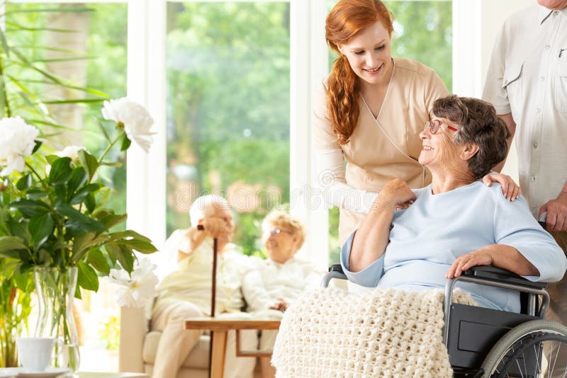 Tender caregiver saying goodbye to an elderly pensioner in a wheelchair in a day care facility. A companion pushing the wheelchair. Other elderly people in the blurred background.