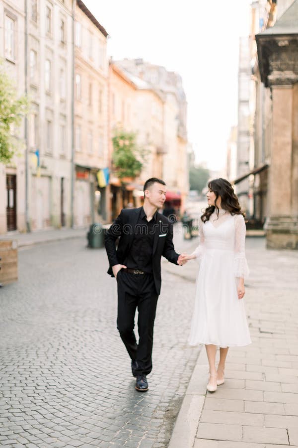 Tender pareja asiática enamorada: novia vestida de blanco y novio con un traje negro en el paseo de su boda en la ciudad vieja foto de archivo Tender pareja asiática enamorada: novia vestida de blanco y novio con un traje negro en el paseo de su boda en la ciudad vieja foto de archivo