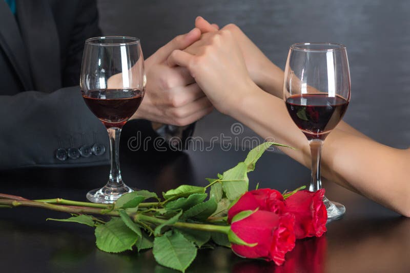 Tenersi per mano dell'uomo della ragazza sulla tavola del ristorante con due vetri del vino rosso e le rose rosse fioriscono fotografie stock libere da diritti Tenersi per mano dell'uomo della ragazza sulla tavola del ristorante con due vetri del vino rosso e le rose rosse fioriscono fotografie stock libere da diritti