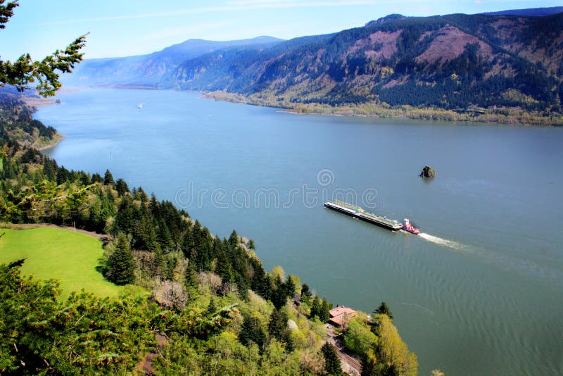 Beautiful majestic territorial view of the Columbia River Gorge with a tugboat pushing some barges. Green trees in forefront, sweeping majestic Columbia River, territorial mountains of the Cascade Mountain Range. Beautiful majestic territorial view of the Columbia River Gorge with a tugboat pushing some barges. Green trees in forefront, sweeping majestic Columbia River, territorial mountains of the Cascade Mountain Range.