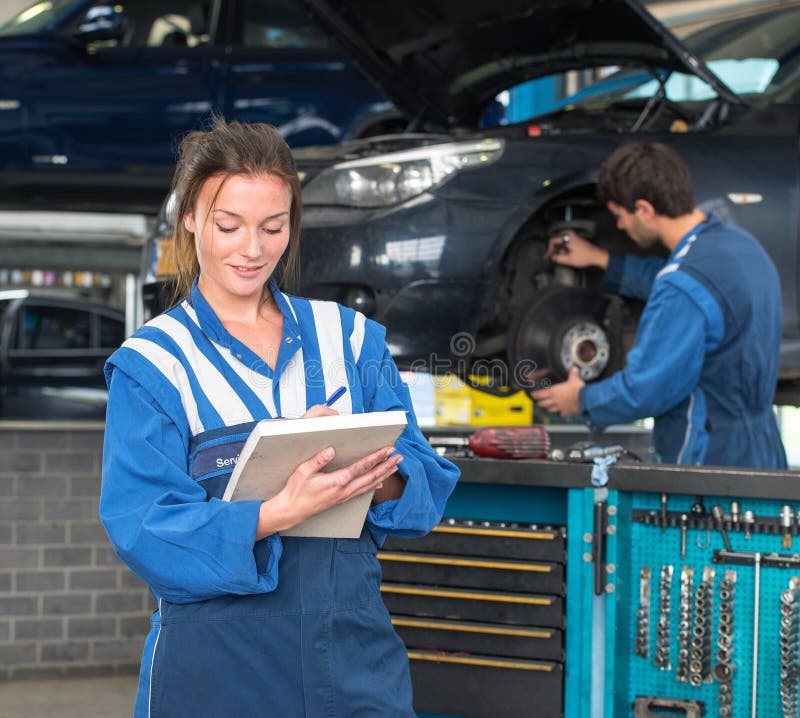 Two mechanics, working in a garage on a MOT test of a car, with a female mechainc in front, checking off inspection items on a note block, with a tool trolley and her co-worker in the background. Two mechanics, working in a garage on a MOT test of a car, with a female mechainc in front, checking off inspection items on a note block, with a tool trolley and her co-worker in the background