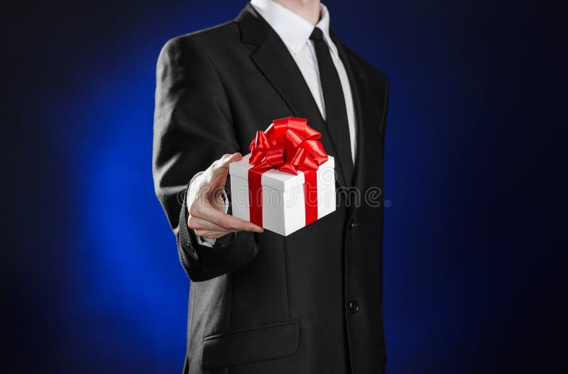 Theme holidays and gifts: a man in a black suit holds an exclusive gift in a white box wrapped with red ribbon and bow on a dark stock photo Theme holidays and gifts: a man in a black suit holds an exclusive gift in a white box wrapped with red ribbon and bow on a dark stock photo