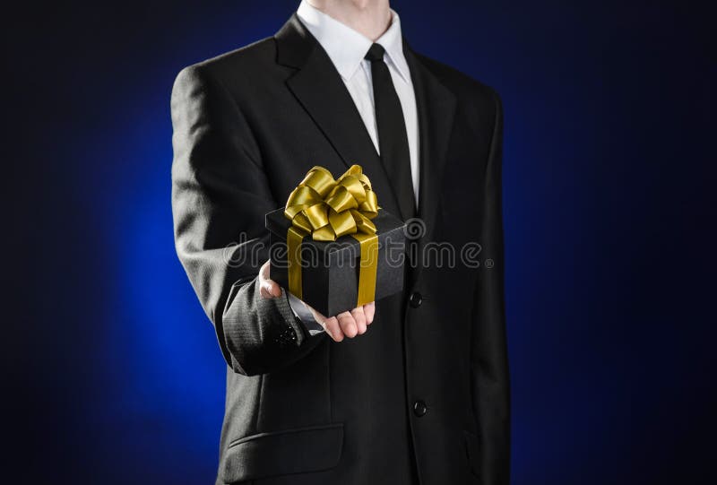 Theme holidays and gifts: a man in a black suit holds exclusive gift wrapped in a black box with gold ribbon and bow on a dark stock photo Theme holidays and gifts: a man in a black suit holds exclusive gift wrapped in a black box with gold ribbon and bow on a dark stock photo
