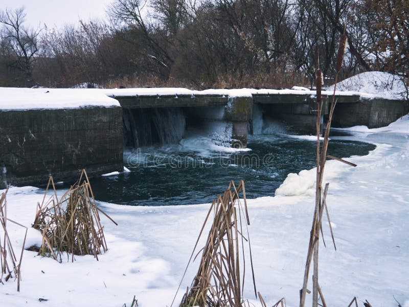 There is a concrete dam on the forest river, ice has frozen on the surface of the water, but water flows from the dam lock royalty free stock photos