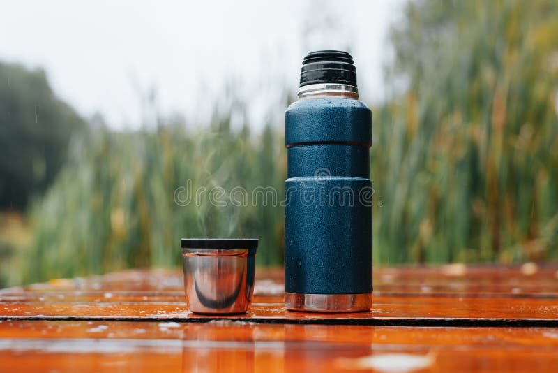 Thermos and Mug with Hot Drink Standing on Wet Wooden Table after