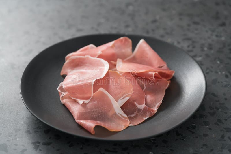 Thin Italian Prosciutto Slices on Black Plate on Conctere Background