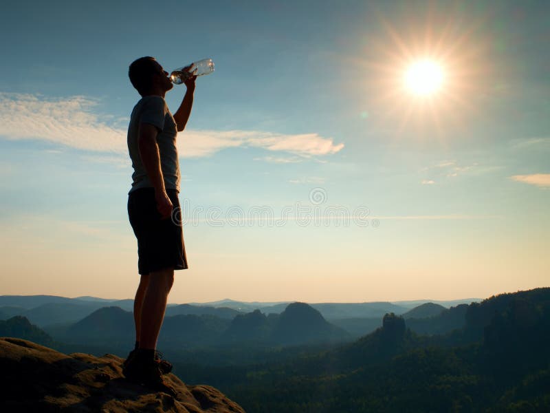 Thirsty hiker in black pants drinks from bottle of water. Sweaty tired tourist on the peak of sandstone rocky park Saxony Switzerl