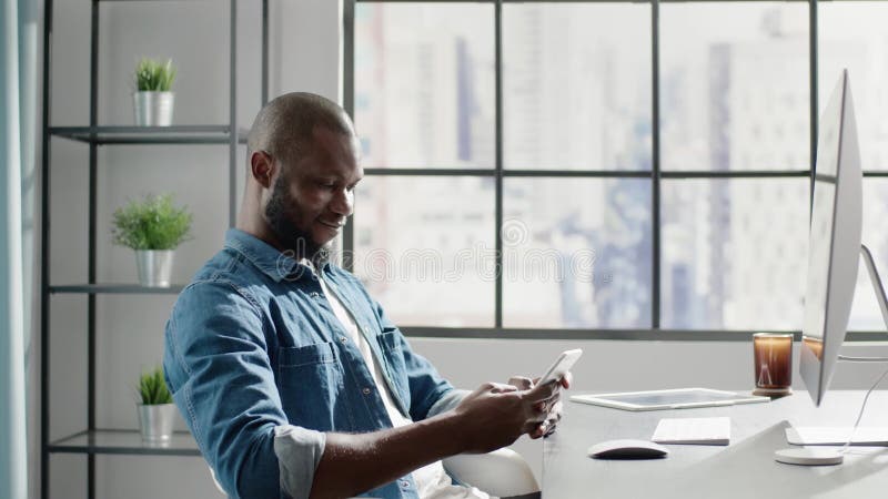 Thoughtful black man in denim chats on smartphone in office