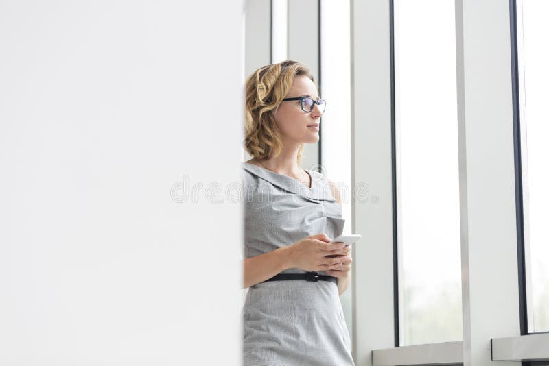 Thoughtful businesswoman holding smartphone while standing by window at new office