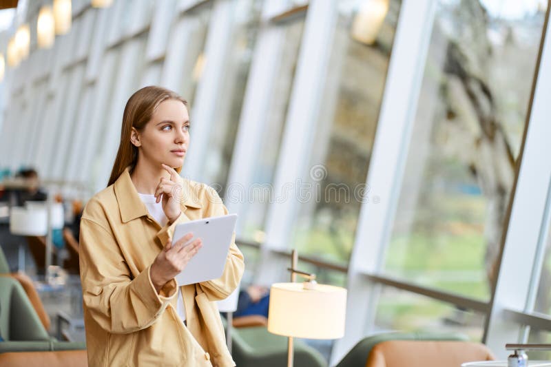 Thoughtful girl student using digital tablet looking away. Copy space. Young woman holding tab computer modern tech device standing in university campus, coworking office space and thinking.