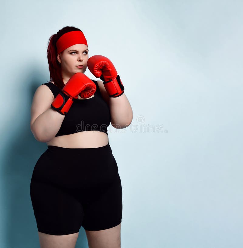 Thoughtful plump  girl in black sporty top, shorts and red boxing gloves looks up thinking over strategy in forthcoming fight.